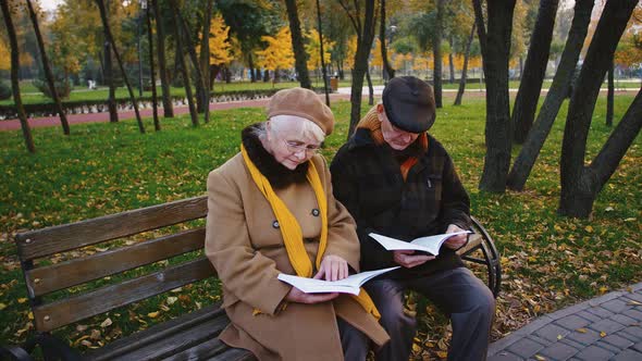 Retired Pair in Glasses and Warm Outerwear are Reading Books and Talking While Sitting on Wooden