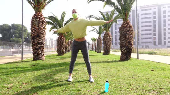 woman with mask exercising on grass in avenue surrounded by palm trees