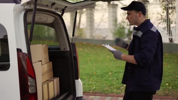 Handsome Courier Making Notes in Documents and Takes Cardboard Box Package Out of Delivery Car
