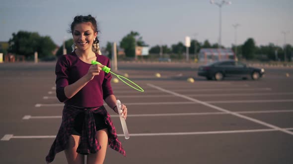 Young Playful Hipster Girl with Long Braids Blowing Bubbles on the Parking By the City Mall During