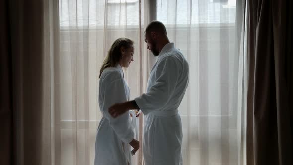 Lovers Dressed White Bathrobes are Standing in Bedroom Against Window