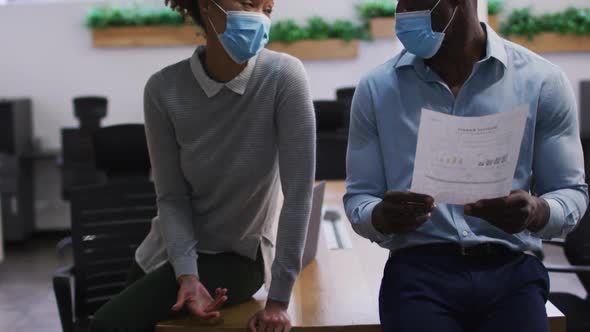 Diverse male and female business colleagues in face masks talking and looking at documents in office