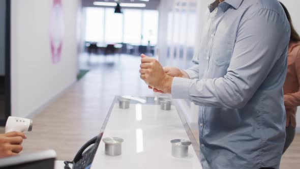 Mixed race man wearing face mask has temperature checked by male colleague at office reception desk