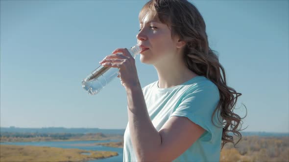 Woman of European Appearance in the Open Air Drinks Water From a Bottle Against a Clear Blue Sky