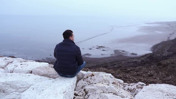 Man in Black Jacket Sits on Large White Stones Near Cold Sea