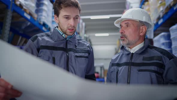 Concentrated Serious Men Examining Paperwork Discussing Plan Standing in Warehouse Indoors