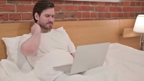 Casual Young Man with Neck Pain Working on Laptop in Bed