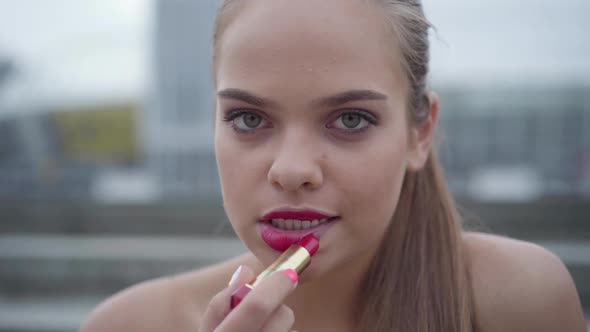 Close-up Portrait of Beautiful Young Girl in Emerald Dress Painting Lips with Bright Pink Lipstick