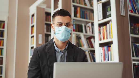 Man in Eyeglasses and Mask Sitting in Library alt