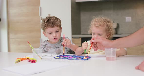 Brother and Sister Draw Watercolors While Sitting at the Table Fathers's Hand Appears with Brush