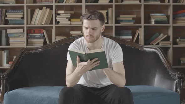 Portrait of Smiling Man Sitting on Couch and Reading Book with Bookshelves at the Background