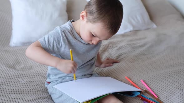 A Small Caucasian Boy in a Gray Tshirt and Shorts on the Bed Draws with Colored Pencils in a