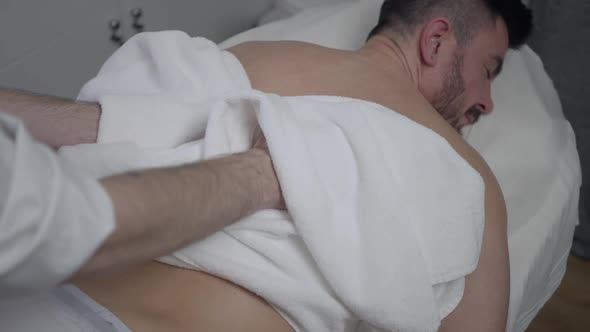 Unrecognizable Masseur Rubbing with Towel Back Skin of Handsome Bearded Man Lying on Massage Bed