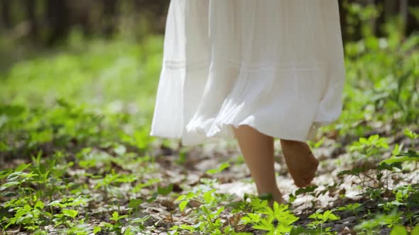 A Woman in a White Dress Walks Barefoot Through the Forest Among Green Plants