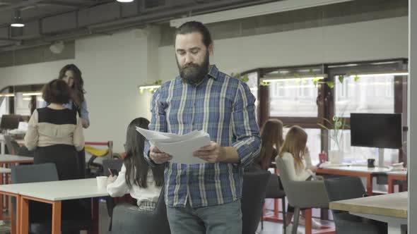 Concentrated Serious Bearded Man Studying Papers Documents Standing in the Modern Office. Two Women