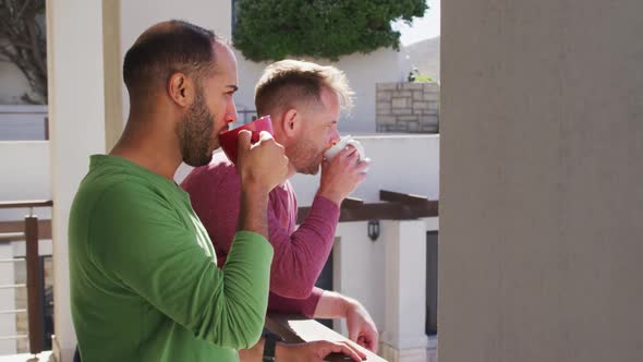 Multi ethnic gay male couple drinking coffee on balcony in sun