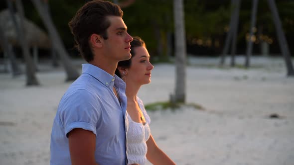 Teenage couple walks on beach during golden hour sunset light. Pretty girl in white dress and boy we