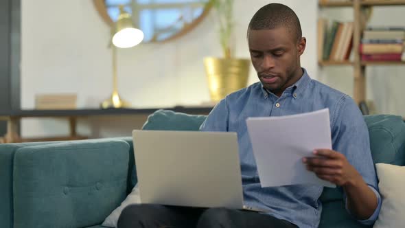 Young African Man with Documents Working on Laptop on Sofa