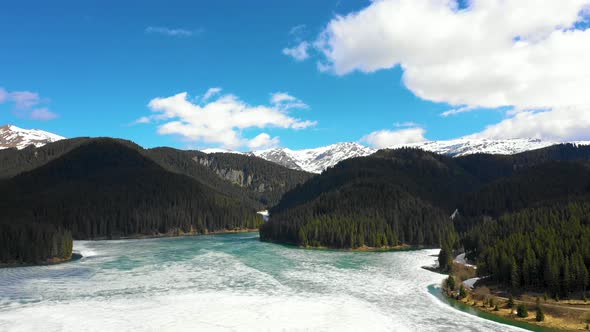 Aerial view of a frozen mountain lake surrounded by forest and snowy mountains in spring season.