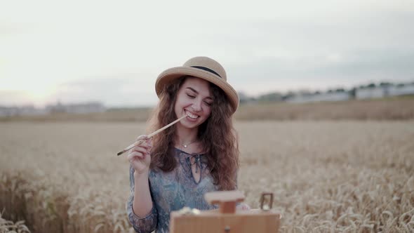 Portrait of Pretty Lady in Hat Poses with Paintbrush and Smiles Among Field
