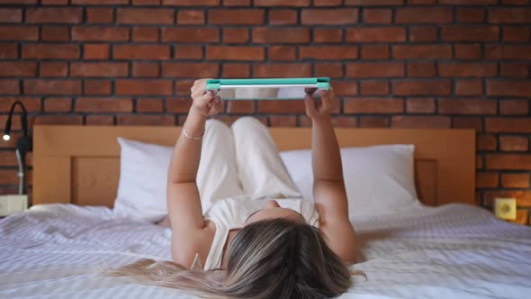Relaxed Young Caucasian Woman Lying on Bed Watching Internet on Digital Tablet Indoors