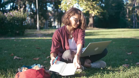 Student Girl Sitting on the Lawn in the Park with Her Laptop and a Notebook