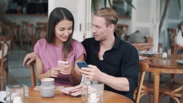 Young Man and Woman Sitting in Cafe, Looking at Smartphone, Laughing and Talking