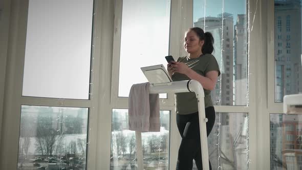 Young female is typing messages on smartphone during walking on treadmill at home