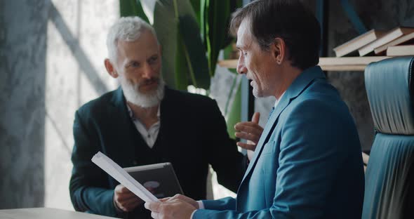 Two Mature Businessmen Discussing Business Growth in Modern Office While Sitting at Desk