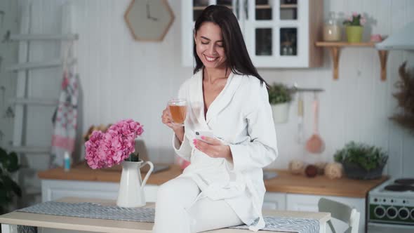 Woman Sits on Kitchen Table, Drinks Tea and Uses Phone for Online Chatting