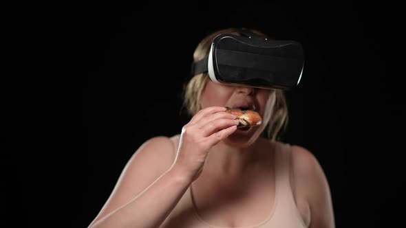 Overweight Caucasian Woman in VR Headset Eating Delicious Unhealthy Bun at Black Background