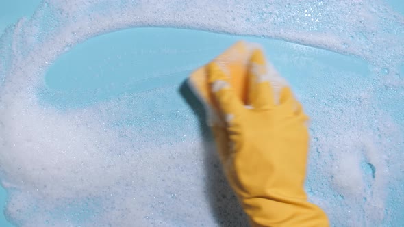 Hand in a Yellow Rubber Glove Holds a Cleaning Sponge and Wipes a Soapy Foam on a Blue Background