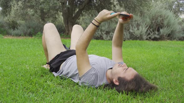 Man with long hair lying on the grass in the park and playing video games