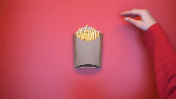 Woman Hand Taking French-Fried Potato From Carton Box on Pink Background, Meal