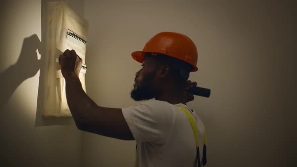 African Electrician with Flashlight Examining Electrical Box with Pliers in New House