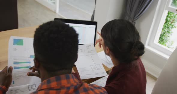 Diverse couple sitting at table talking and working with laptop