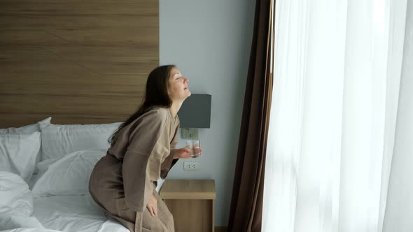Happy Lady in Bathrobe Drinks Water By Hotel Room Window