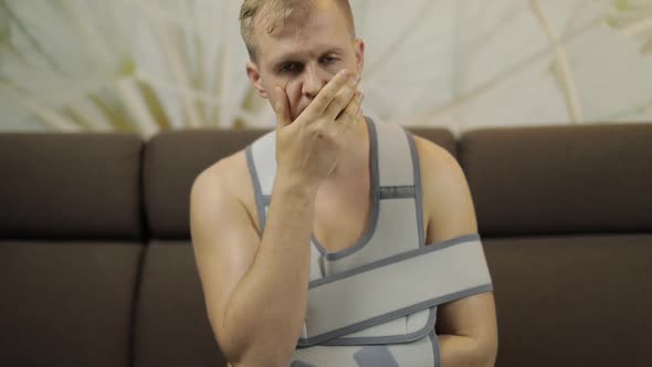 Painful, Bored Man with a Broken Arm Wearing Arm Brace Sitting on a Sofa