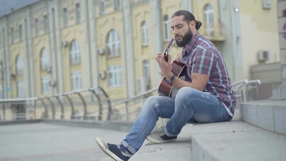 Wide Shot Side View of Young Caucasian Man Sitting on Urban Stairs and Playing Guitar. Handsome
