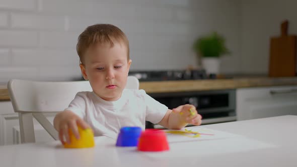 Cute Little Baby Boy Sitting Behind Table and Drawing with Fingers Covered in Colorufl Paint