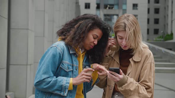 Two Diverse Young Women Smiling and Use Smartphone While Standing on the Street