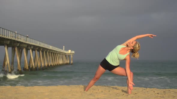 A young woman does yoga on the beach next to a pier.