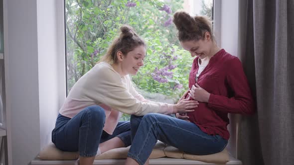 Side View Excited Young Woman Admiring Caressing Belly of Pregnant Twin Sister Sitting on Windowsill