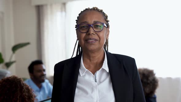 African senior woman looking on camera inside bank office - Business people working in background