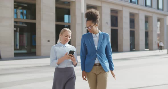 Tall and Short Diverse Women in Office Clothes Walking Outdoors and Using Phone