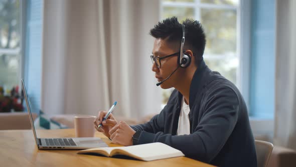 Asian Male Student in Headset Watching Online Webinar on Laptop and Writing in Notebook at Home