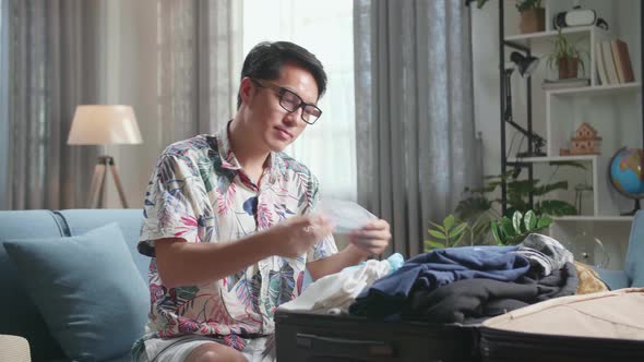 Asian Young Man Packing Clothes And Protect Face Mask In Suitcase At Home, Preparing For Vacation