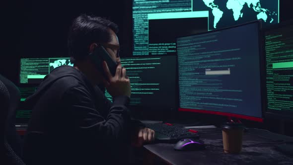 Asian Male Hacker Talking On Phone With Multiple Computer Screens And Success Downloading Data