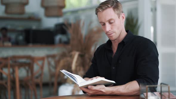 Portrait of Focused Man in Black Shirt Sits in Modern Outdoor Cafe, Reads Book