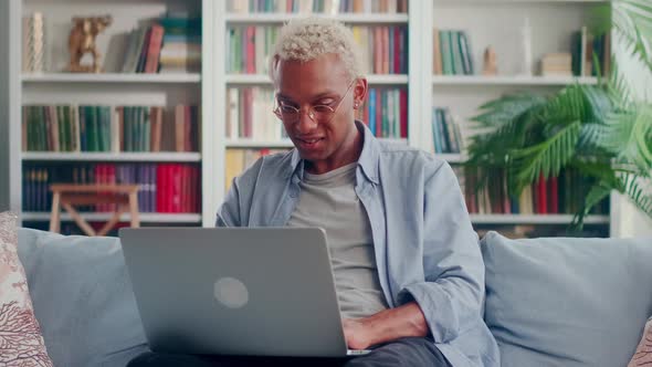 Happy Dark Skinned Man Sitting on Couch Using Laptop Screaming Yes Raising Fist